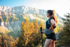 A pregnant woman stands mid hike and gazes out at scenery while holding walking sticks