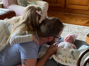 Man in gray shirt laying on floor with daughter on his back looking at infant with a pink hat on