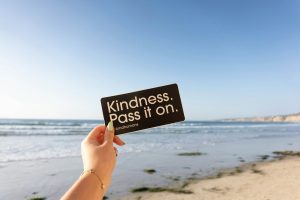 Person holding a note that says "kindness. pass it on" On the beach
